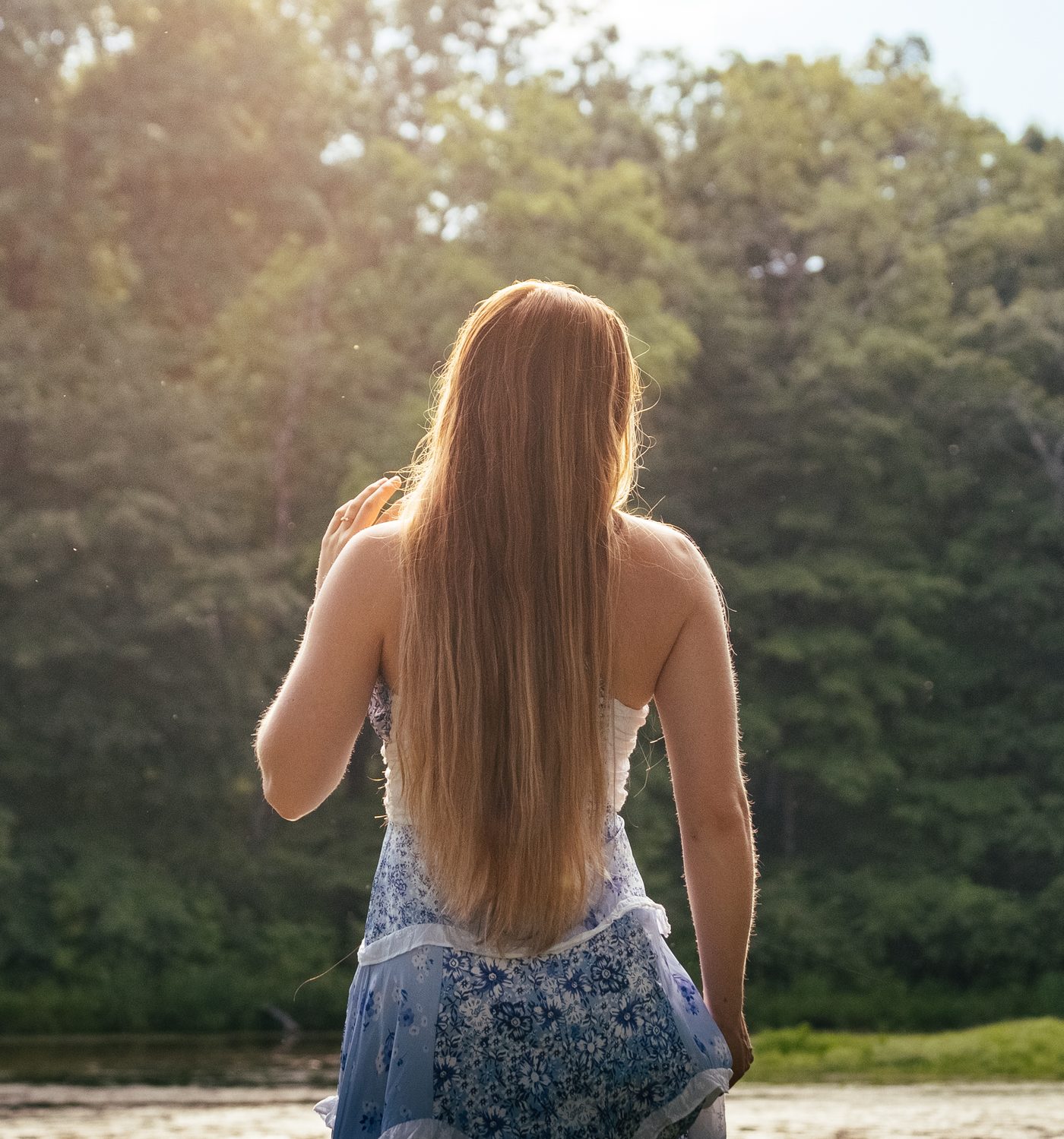 Back view of a woman with long sunlit hair facing a wooded pond
