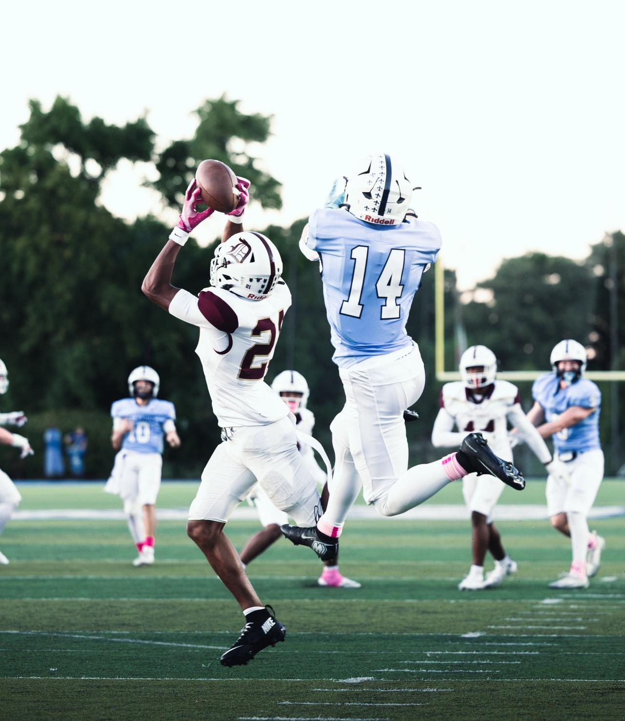 Receiver in white and maroon leaping for a pass beside a defender in light blue