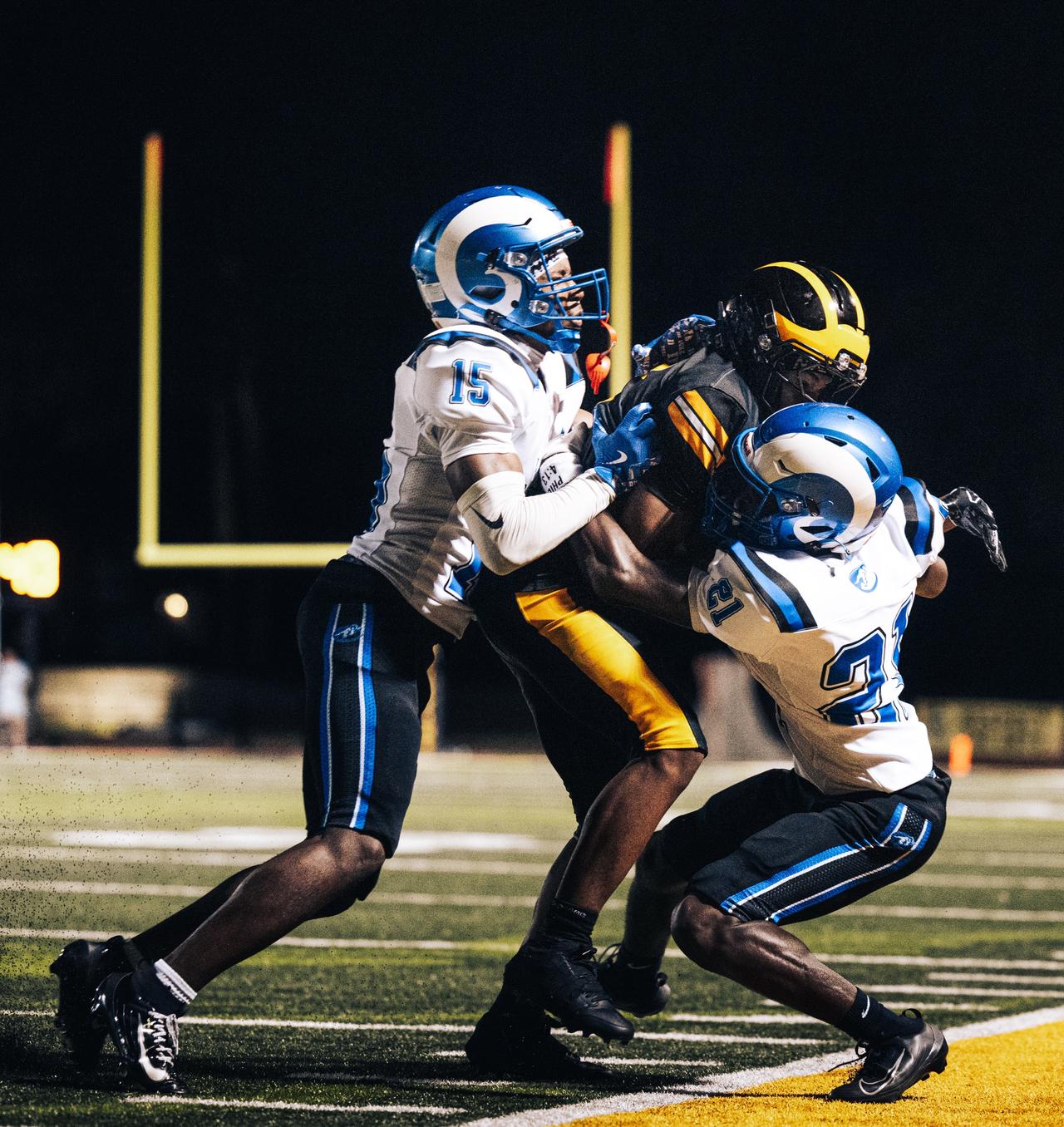 Two blue and white defenders tackling a black and gold ball carrier in front of yellow goalposts