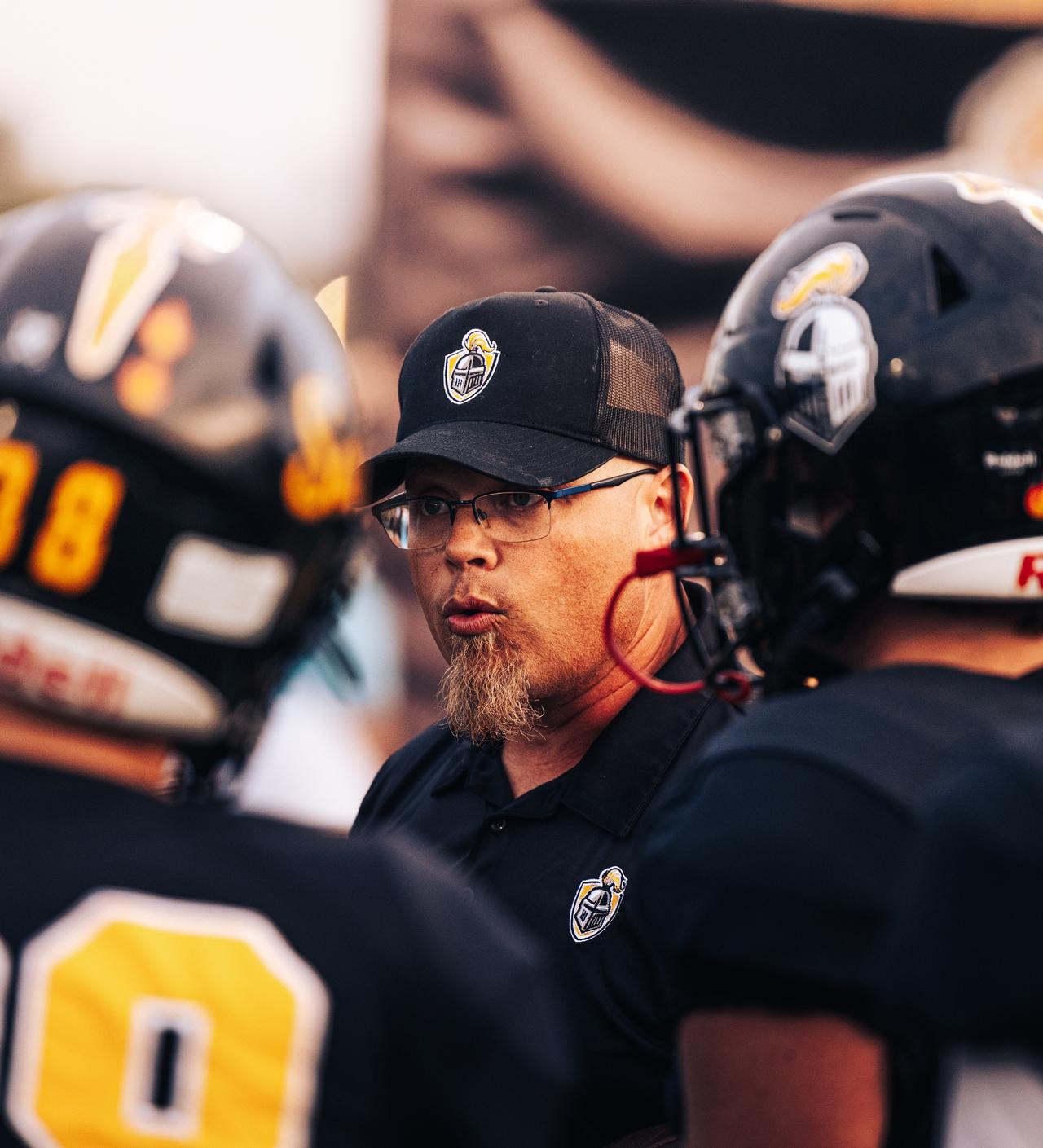 Coach in a black cap and glasses speaking between two blurred helmets